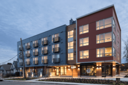 The Jefferson Station apartments at dusk. The left portion of the building shows the apartments' balconies while the right side features very large windows. The bottom floor has a wood slat exterior and a corner space for retail.