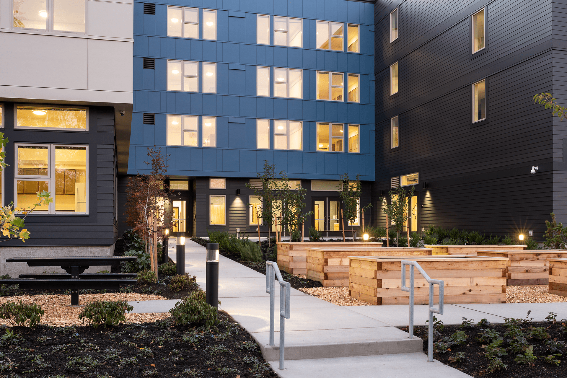 An exterior photo of Kent Supportive Housing hows its white, blue, and gray siding, as well the wood planter boxes leading to the entry.