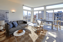 The interior of one of the units in the Lexicon Apartment building includes gray wood floors, a seating area, dining table, and large corner windows.