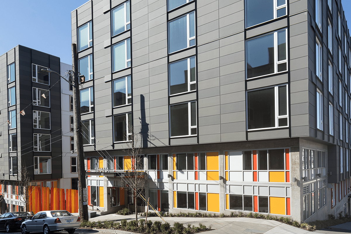 The exterior of the Hana Place building has four floors with gray siding and a colorful ground floor with an orange, yellow, and white exterior.