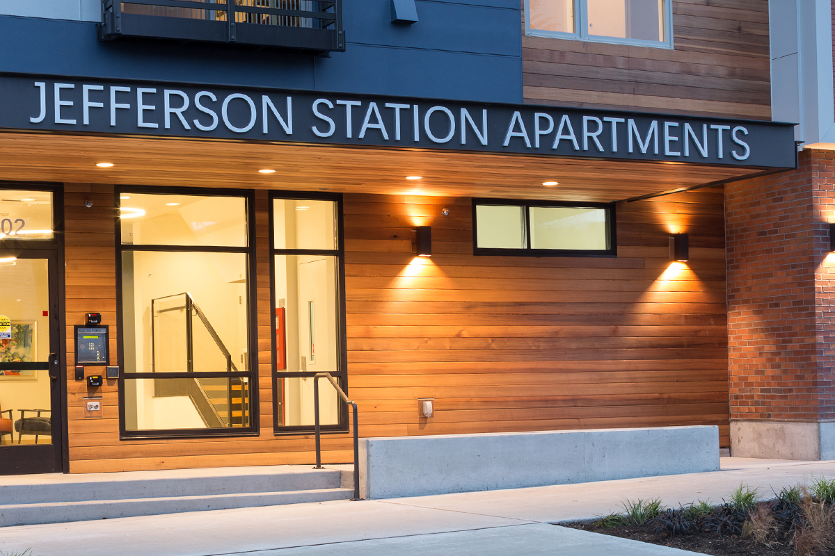 A close shot of the entry to the Jefferson Station apartments shows its wood slat exterior, tall black-trimmed windows, the front door, and a sidewalk.